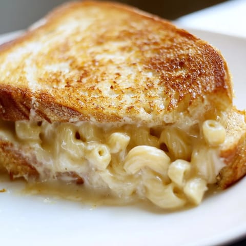 A close-up view of a hearty Mac & Cheese Grilled Cheese, cut diagonally to reveal layers of gooey, comforting pasta tucked between crispy buttered bread.